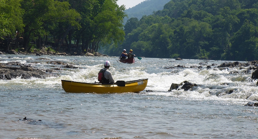 In the background, two students paddle a canoe toward whitewater. In the foreground, another person, likely an instructor, watches them from a canoe.